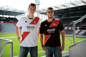 Dennis Daube und Michael Gregoritsch präsentieren das neue Trikot, nun mit Sponsorenaufdruck. Sieht für mich aus der Ferne wie ein auf der Brust getragenes Arschgeweih aus. Foto: coca-cola-deutschland.de