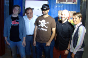 Vier Männer und eine Frau vor einer blauen Bretterwand mit FC St. Pauli-Ausstellungsstücken. Der in der Mitte stehende, blinde Mann trägt eine Mixed-Reality-Brille